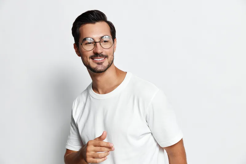 Smiling man wearing glasses and a white t-shirt poses confidently while pointing with his finger, showcasing a vibrant personality against a plain light background.