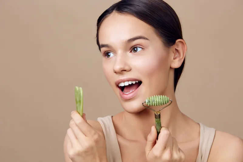 Young woman using jade roller and cucumber slices for skincare, showcasing her enthusiasm for beauty treatments and healthy skin, against a neutral background