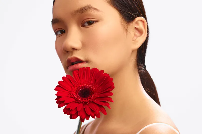 Asian woman holds a vibrant red flower near her face, expressing calm confidence and subtle beauty in a minimal studio setting with soft lighting and a clean background.