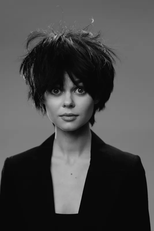 Black and white portrait of a woman with messy hair wearing a dark blazer looking at the camera