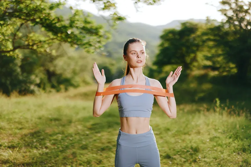 Young athletic woman exercising outdoors with resistance bands in a natural setting, showcasing strength and determination during a fitness routine