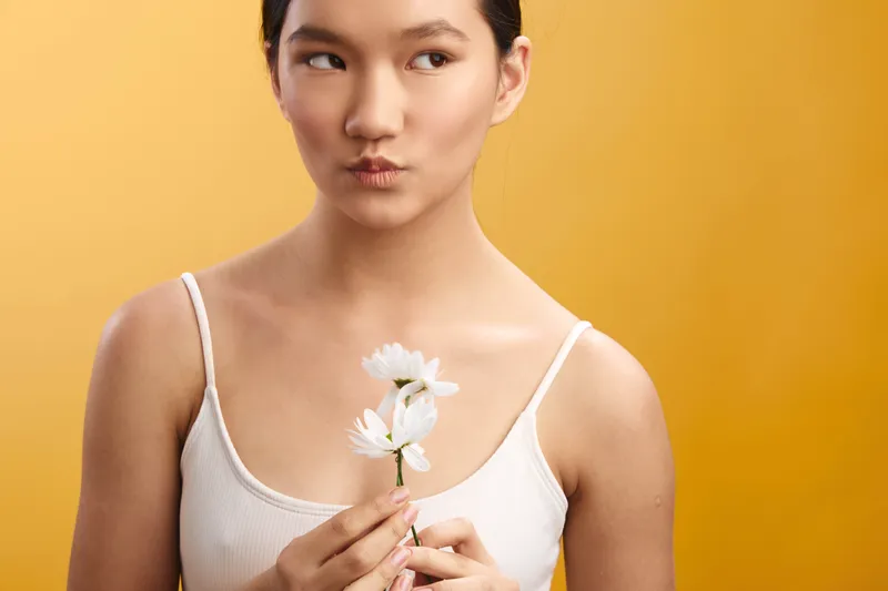 asian woman wearing a light top stands before a warm orange backdrop, holding a white flower gently in front of her, with a soft pout and subtle smile, conveying calm charm.