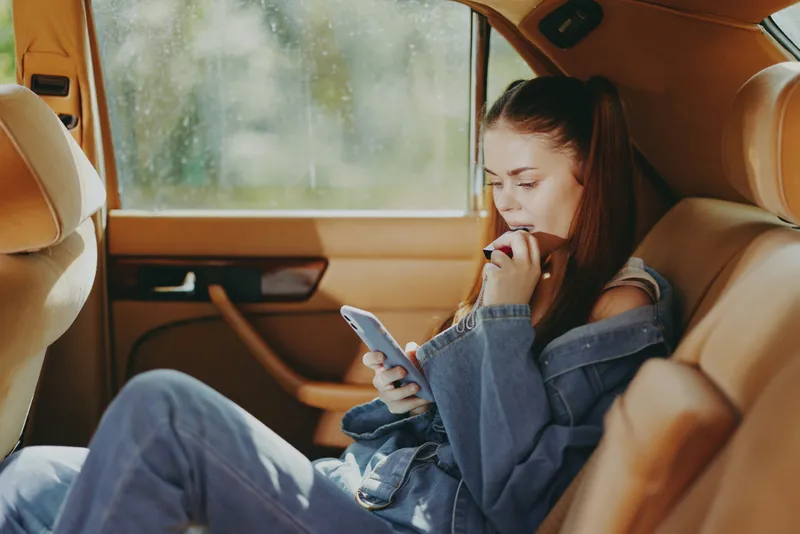 Young woman with long hair using a smartphone in a vintage car, wearing denim jacket, thoughtful expression, relaxed atmosphere, natural light, casual style.