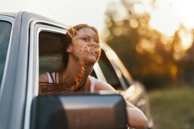 Young woman enjoying a sunset drive, hair blowing in the wind, with a cheerful expression, showcasing freedom and adventure, in a silver car.