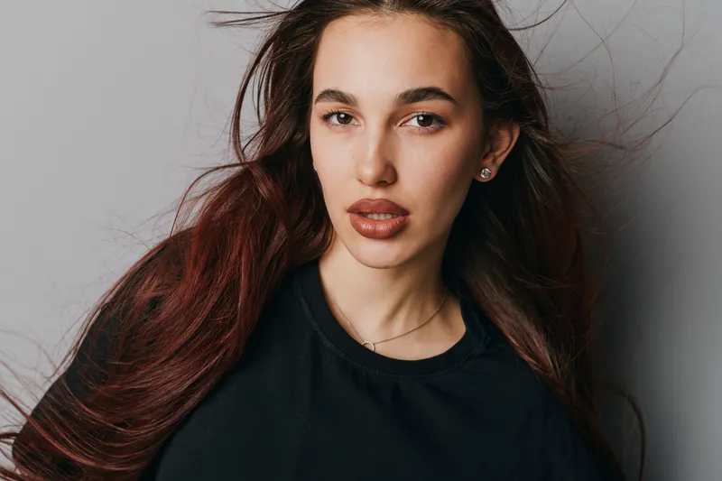 Woman with long hair and natural makeup posing against gray wall, wearing black shirt, looking at camera with relaxed expression and soft lighting on face and hair.
