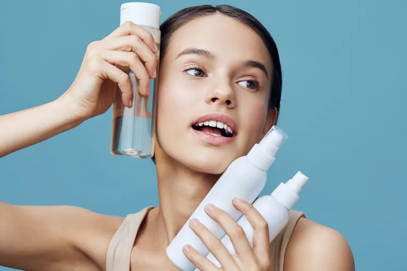 Young woman showcasing skincare products with a fresh, natural look against a light blue background, expressing joy and confidence in her skincare routine