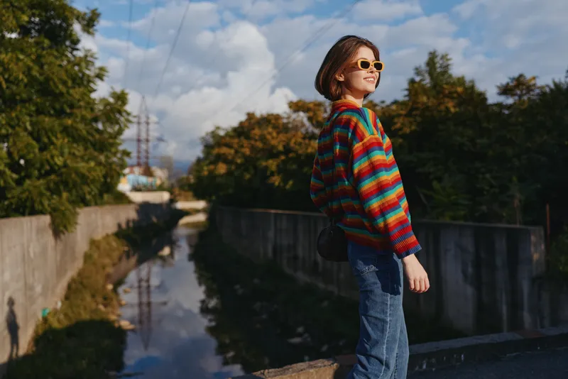 Woman in a colorful striped sweater and sunglasses stands on a concrete bridge by a canal, smiling and looking sideways in a casual outfit, short hair, sunny urban travel lifestyle scene.