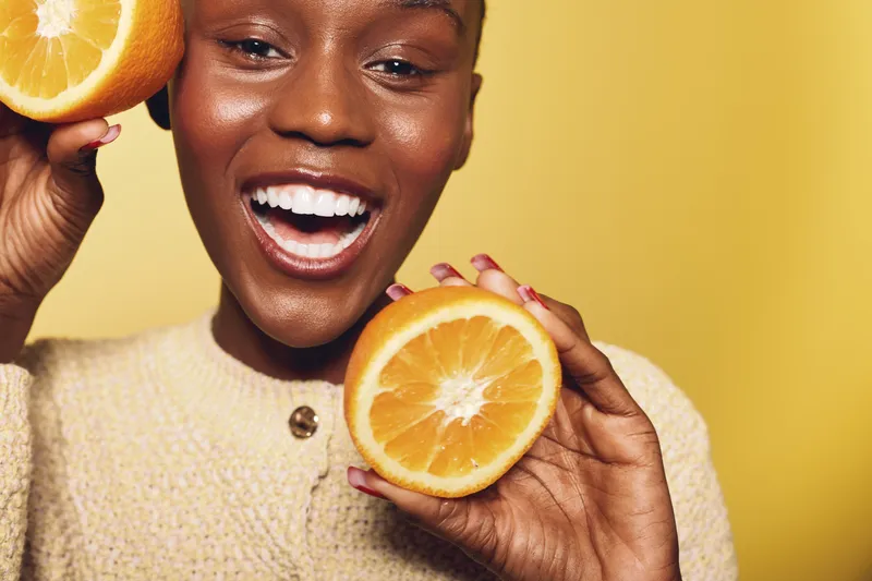 Smiling black woman with dark skin holding two halves of a fresh orange fruit close to her face on yellow background. Concept of healthy lifestyle, vitamin C, and natural beauty.