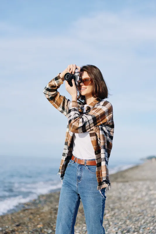 Photographer woman on a rocky seaside shooting with a camera, wearing plaid shirt and jeans, enjoying a windy day by the ocean with bright sky and calm water in the background