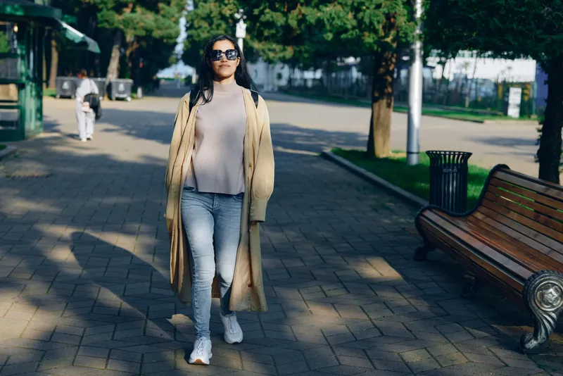 Woman walking along a park street in a beige long coat, casual chic style with jeans and white sneakers, sunglasses, daytime urban scene and relaxed confidence