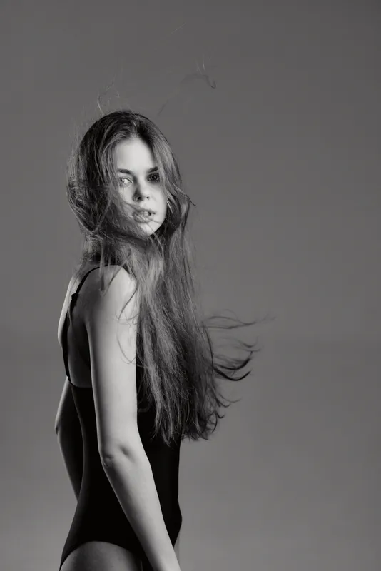 Black and white portrait of a young woman with long hair in a black outfit, looking over her shoulder with a calm expression.