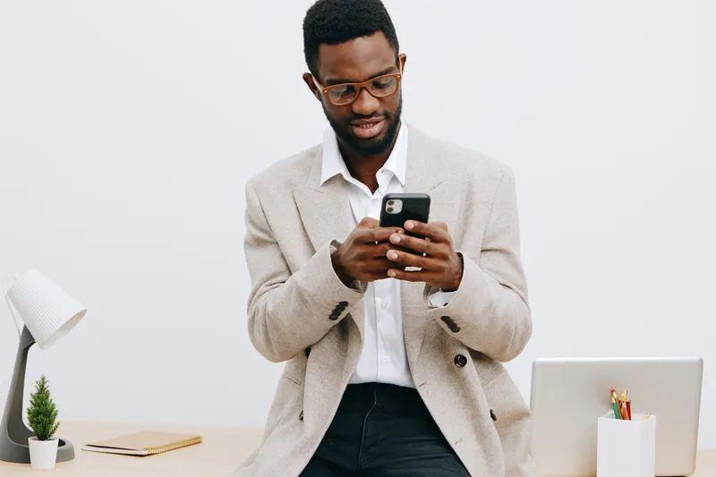 Professional young man using smartphone in modern office setting, dressed in smart casual beige blazer and white shirt, with laptop and stationery on desk, isolated on plain white background. Business