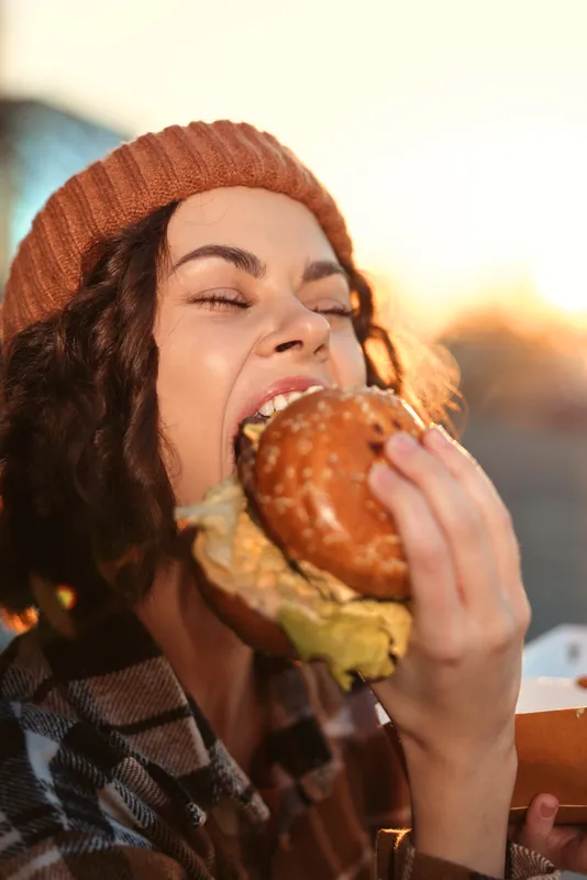 Woman bites into a juicy burger outdoors at golden hour, candid smile and authentic moment of candid lifestyle, authenticity, mindful living and emotional storytelling in warm glow.