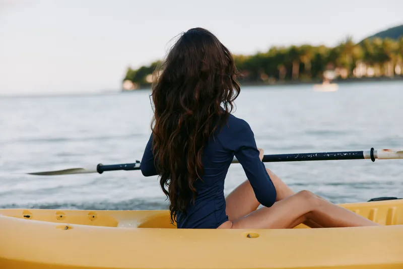A lone figure with long dark hair sits in a yellow rowing boat, facing tranquil water and a distant shoreline, holding an oar as soft light warms the scene.