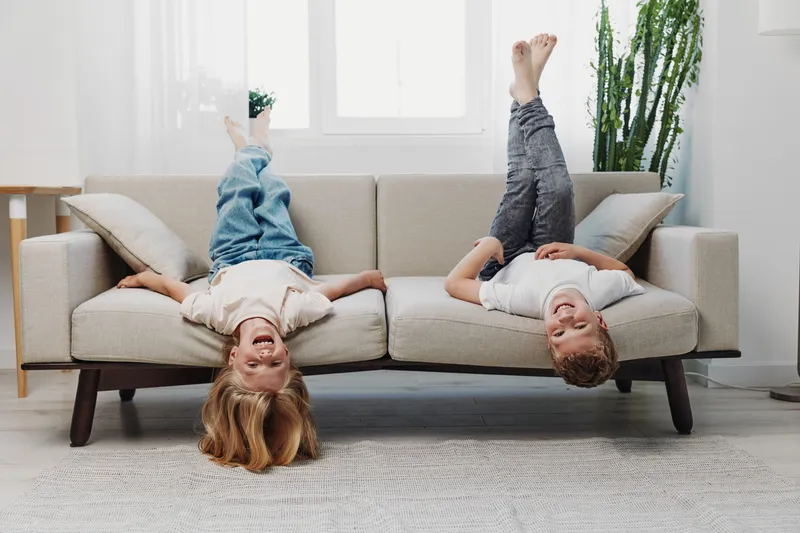Two happy children lie upside down on a beige sofa in a bright living room, smiling and having fun. Concept of playful childhood moments, family joy, and relaxed home atmosphere.