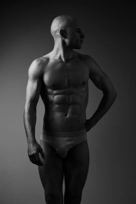 Athletic man posing in black and white lighting, showcasing strength and confidence, highlighting his muscular body and strong profile in a monochromatic setting.