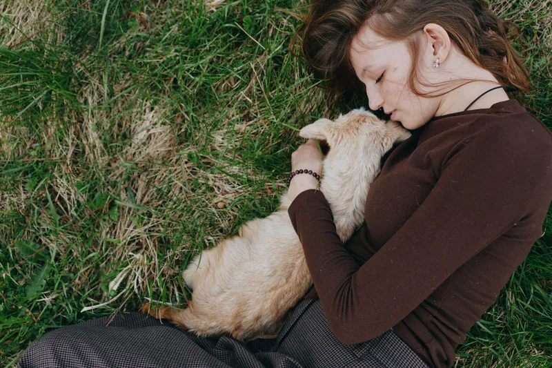 A woman lies on green grass, cuddling a goat kid with a tender smile, sharing a warm, intimate moment outdoors, highlighting rural lifestyle, nature, and gentle human animal bond.