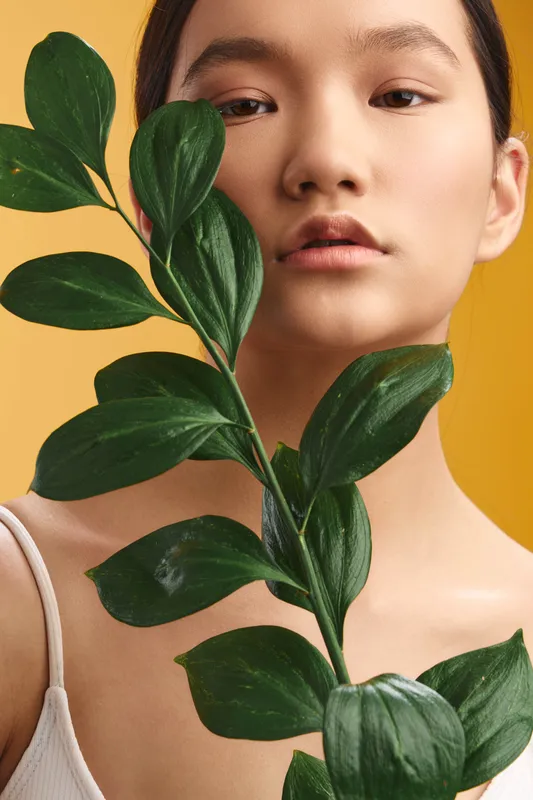 Asian woman poses with a leafy plant against a warm background, presenting a calm, minimalist portrait that emphasizes natural beauty, balance, and gentle elegance.