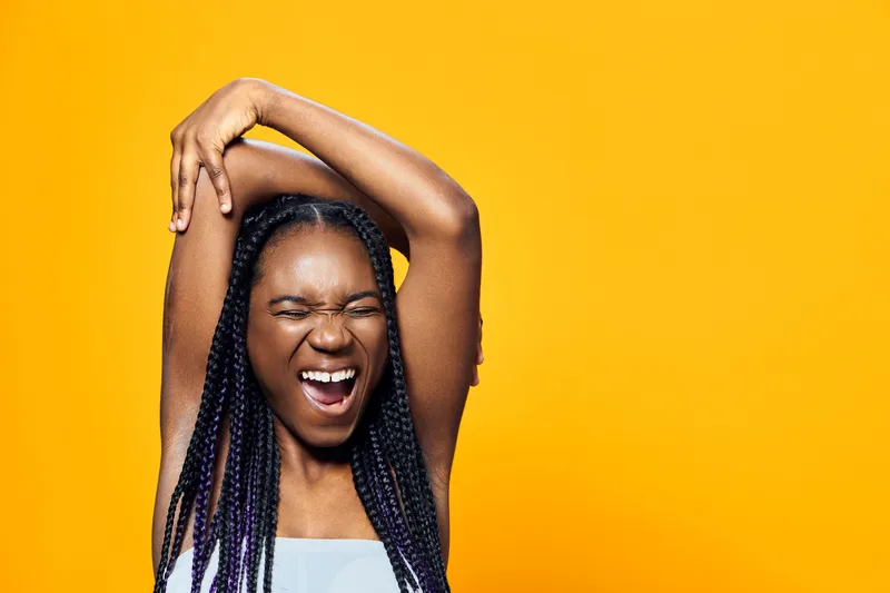 Happy young Black woman with braided hair expressing joy against a bright yellow background, showcasing vibrant colors and cheerful emotions