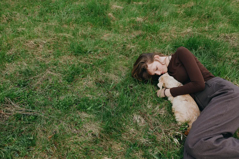 Lifestyle scene with a woman lying on grass, cuddling a dog outdoors, sunlight sparkling through leaves, relaxed pose, peaceful mood, natural colors and greenery creating tranquility throughout a