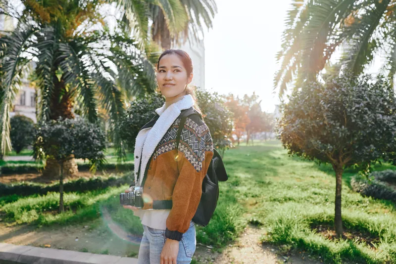Woman with camera in sunny park, outdoor fashion, backpack, casual outfit, capturing moments amid palm trees and green lawn, bright warmth and relaxed mood