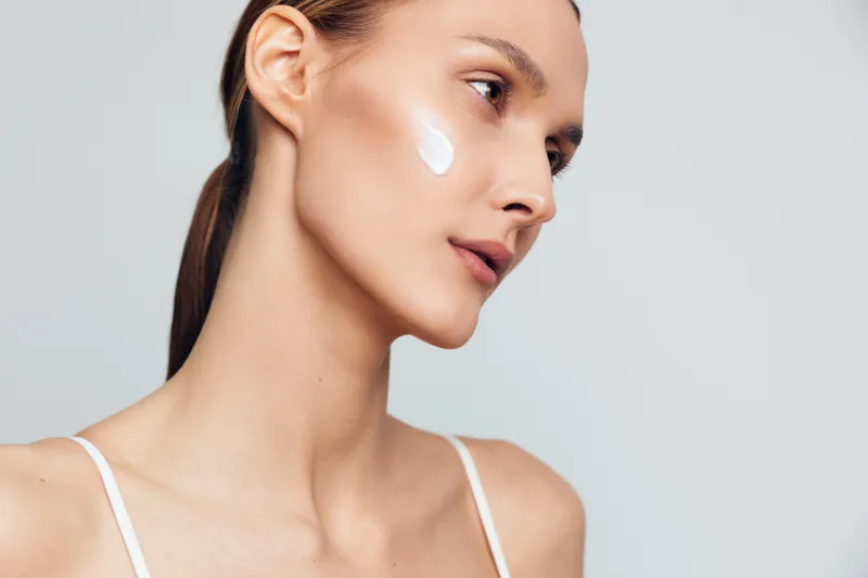 Young woman applying skincare cream on face in profile view, smooth skin and calm expression, studio beauty portrait, isolated on light background, close up