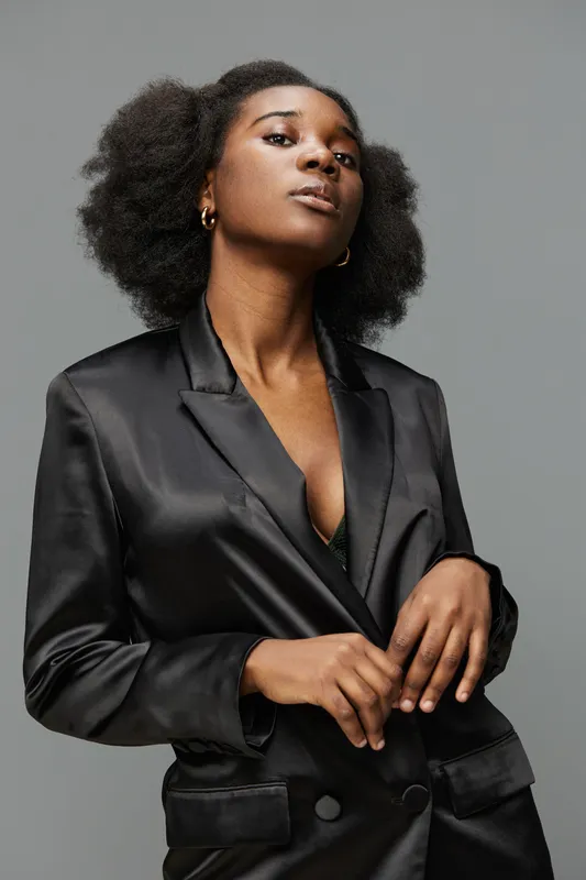 Confident young Black woman in a stylish black satin blazer poses with a bold expression against a neutral grey background