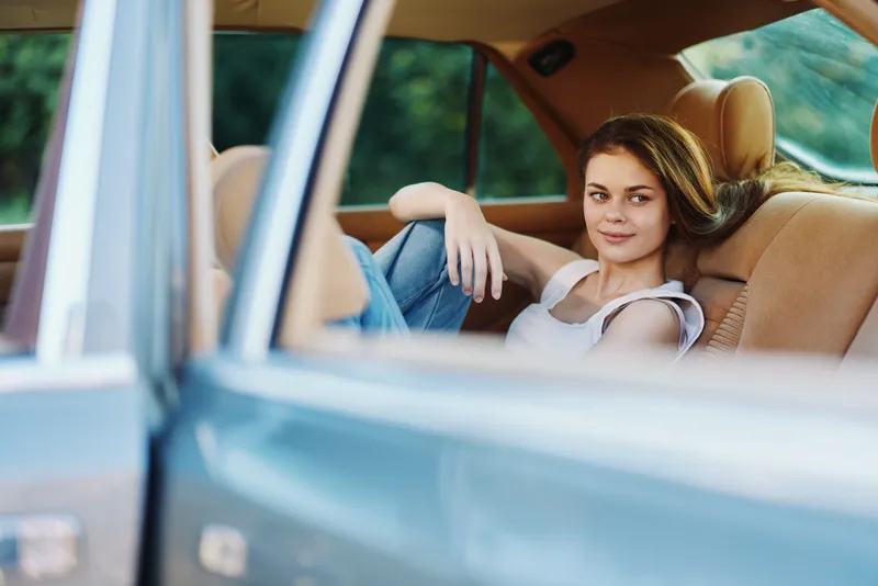 Young woman in casual attire relaxing in a vintage car, exuding confidence and comfort, surrounded by greenery, perfect for lifestyle and travel themes.