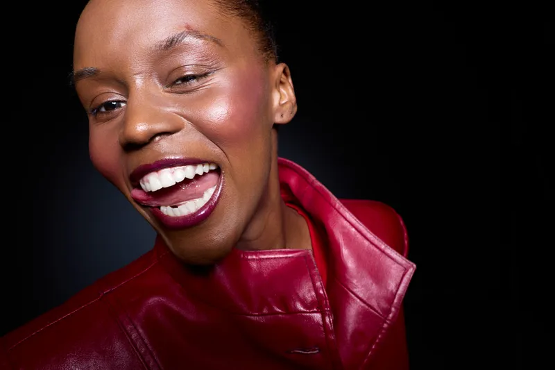 Close-up of happy African woman with dark skin laughing, wearing red leather jacket on black background. Concept of joy, fashion, and confident female portrait in studio lighting.