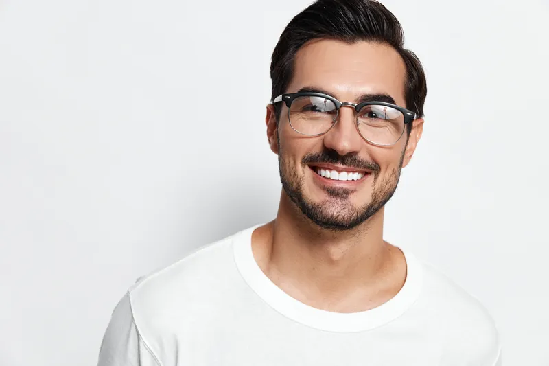 Smiling man with glasses wearing a white t-shirt poses cheerfully while looking at the camera against a solid white background, showcasing a vibrant lifestyle concept.
