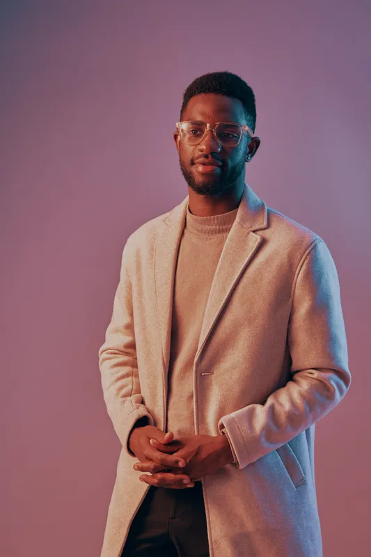 Stylish young man in a light coat with glasses, expressing confidence and elegance against a vibrant, colorful background