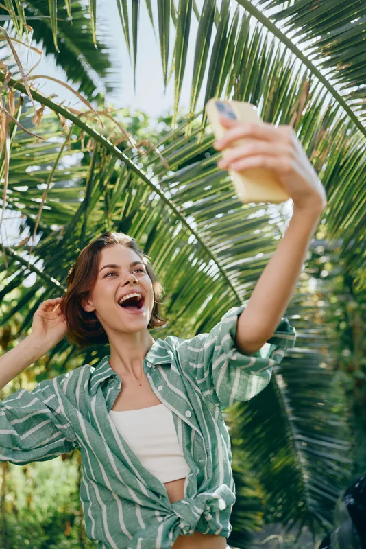 Woman smiling outdoors, joyful moment captured as she lifts a drink in a sunny tropical setting, palm trees overhead, casual attire, carefree mood, vibrant summer atmosphere.