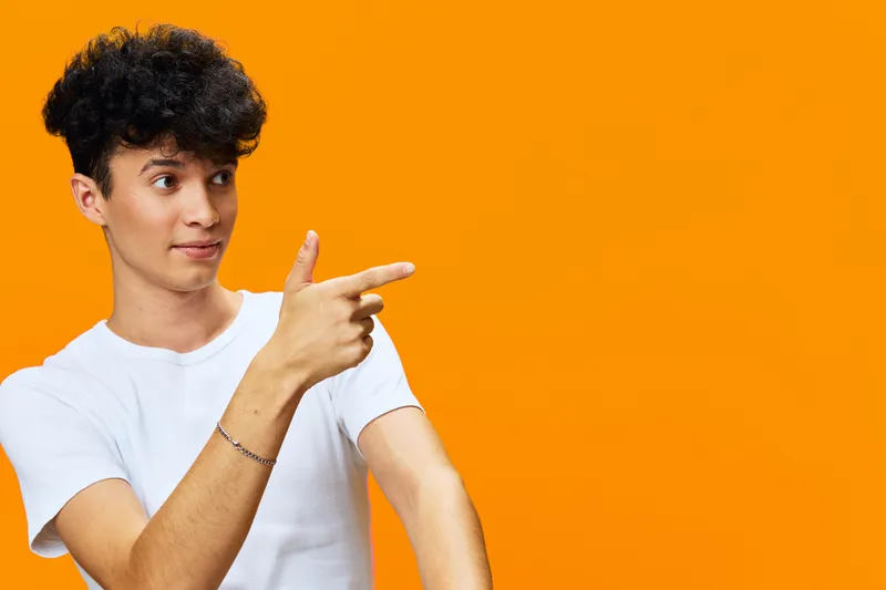 young man pointing finger with curiosity, wearing white t-shirt, short curly hair, expressive face, bright orange background, vibrant lifestyle concept