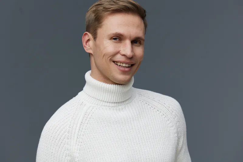 Smiling young man wearing white turtleneck sweater against plain gray background. Male model with cheerful expression, casual clothing, studio portrait, lifestyle concept, winter fashion, happy mood, 