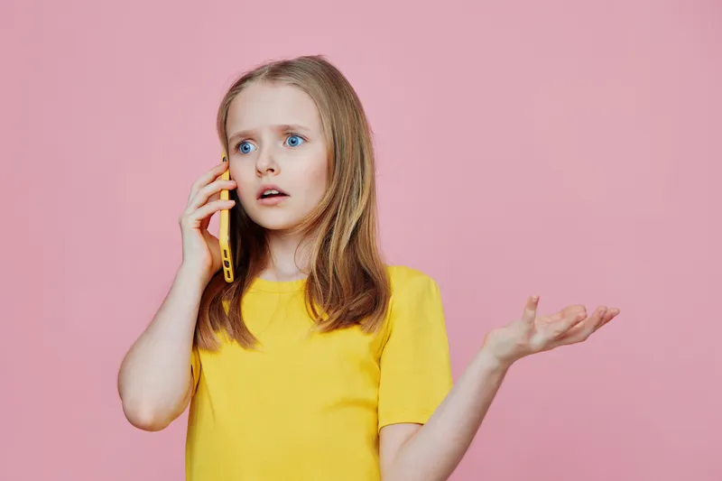 Young girl in a yellow shirt expressing surprise while talking on a phone against a pink background, demonstrating emotions of confusion and curiosity
