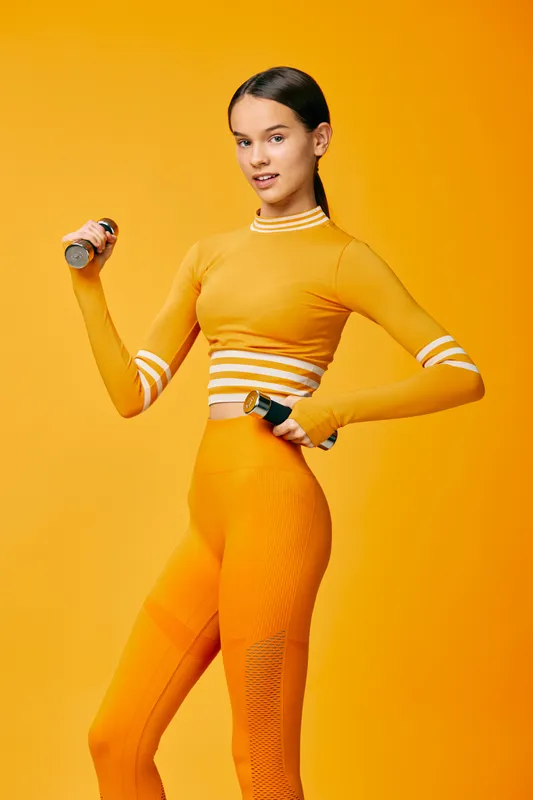 Woman in bright orange fitness outfit lifts dumbbells in a dynamic workout pose, vibrant studio background, emphasizing health, strength and active lifestyle.