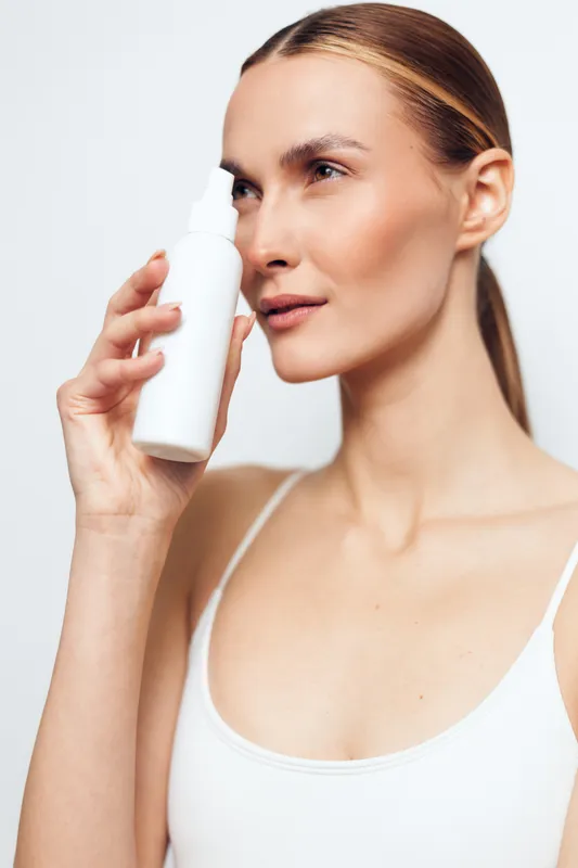Woman with skincare bottle, close up face portrait, clean background, beauty routine using serum, spa wellness mood, young adult female with white bottle, hydration and care, calm expression, looking