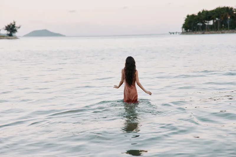 A lone woman stands waist deep in calm sea at dusk, pink dress flowing as gentle waves lap her, distant islands and shoreline form a serene horizon.