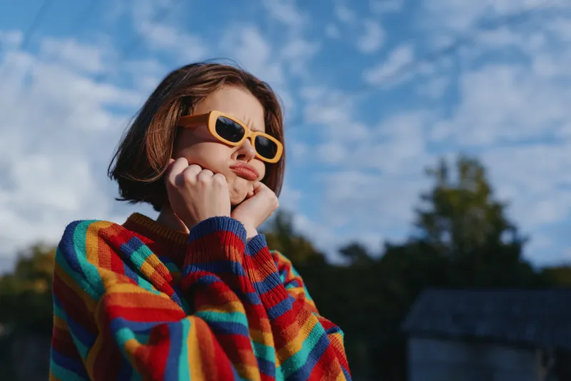 Teen girl (age 13) in a colorful sweater and yellow sunglasses, outdoor portrait under cloudy sky, posing with playful pout and hands on cheeks for casual lifestyle travel fashion.