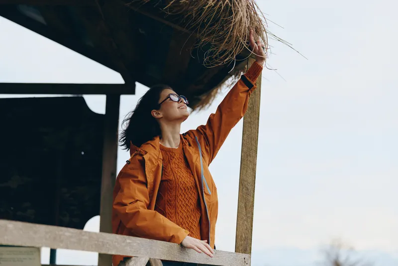 Woman lifestyle portrait outdoors, orange jacket leaning on wooden railing, looking upward in warm sunlight, relaxed casual pose, soft sky background, daylight mood, fresh air freedom