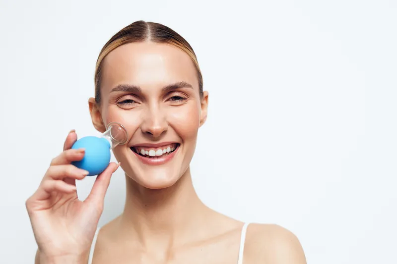 Smiling woman with clear skin holding a blue facial cleansing brush, applying skincare in a bright studio. Concept of beauty, self-care, and healthy skin routine, natural makeup, wellness.
