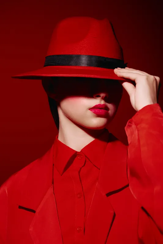 Stylish woman wearing a red hat and matching coat poses against a red background. Her lips are glossy and brightly colored promoting fashion, beauty, and creative style.