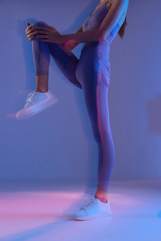 Woman sport fitness wearing purple activewear stretching leg indoors with blue and pink light, demonstrating flexibility and strength in a modern workout setting.