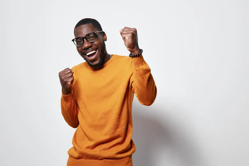African American man with dark skin wearing glasses and orange sweater smiling and celebrating with fists up on white background. Happy young male portrait with excitement.