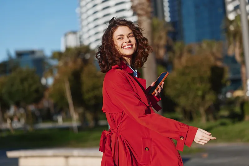 Smiling woman in a red coat outdoors, holding a smartphone. Sunny park setting with urban buildings in the distance. Casual fashion lifestyle scene.