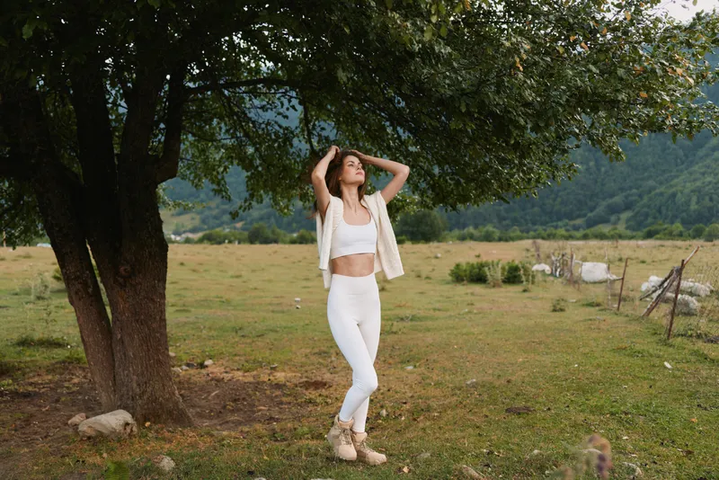 Young woman in white clothes stands under a large tree in a rural field with mountains in the background, enjoying a peaceful moment with her hands in her hair.
