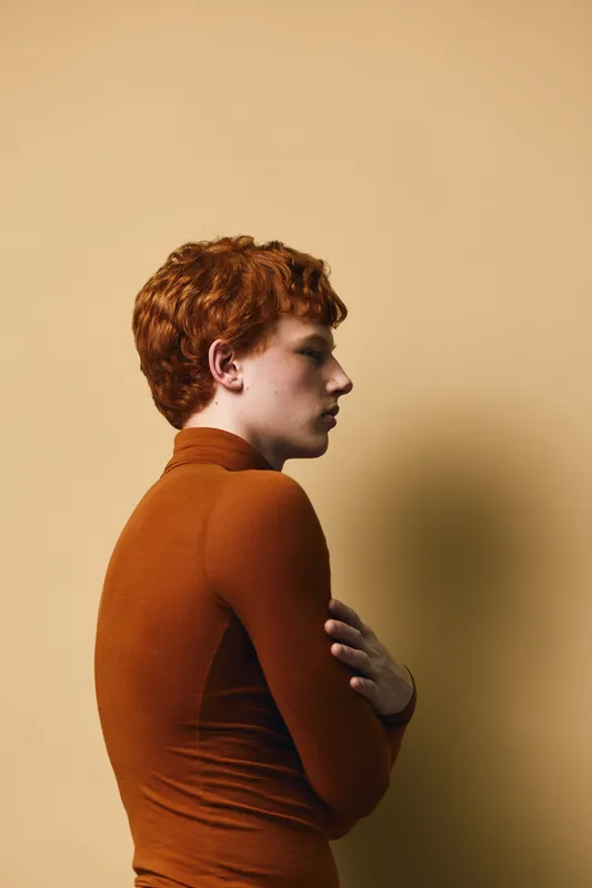 Young redhead man in brown turtleneck sweater standing sideways with crossed arms on beige background, serious expression and casual style portrait indoors.