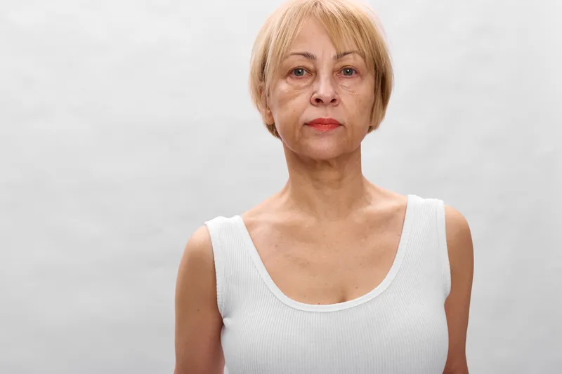 Elderly woman portrait in studio, mature senior female, short blond hair, neutral expression, white sleeveless shirt, plain white background, calm mood