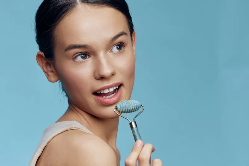 Young woman using a jade roller for facial massage, showcasing natural beauty and skincare routine in a serene blue background