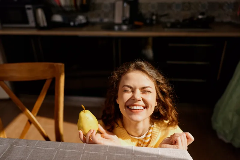 A cheerful woman in a yellow striped top smiles at the camera while offering a pear on a kitchen table, conveying warm, casual home lifestyle. A natural candid moment of comfort. Now.